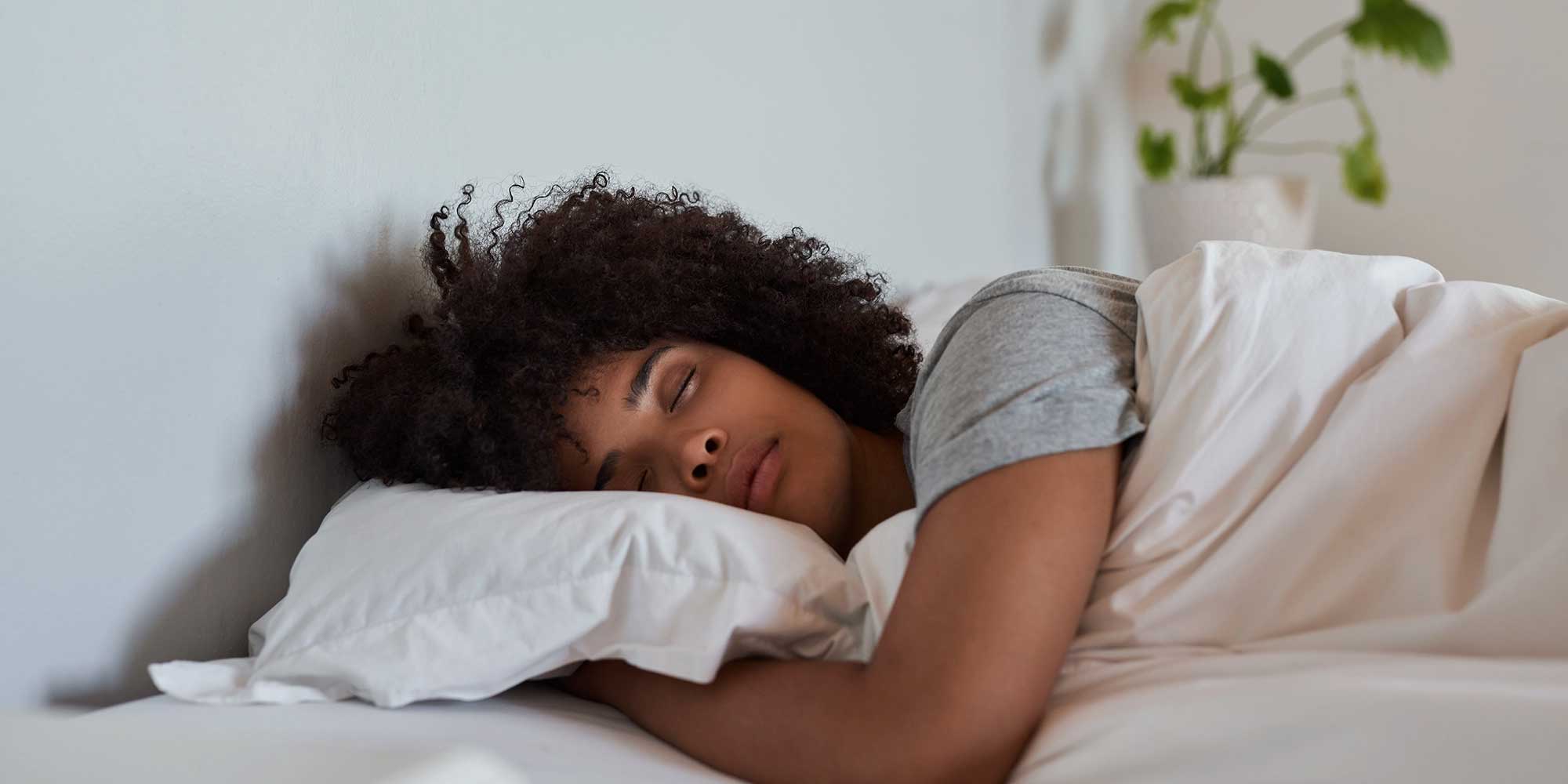 A young woman sleeps peacefully in bed. The bed has white sheets and there is a green plant in the background.