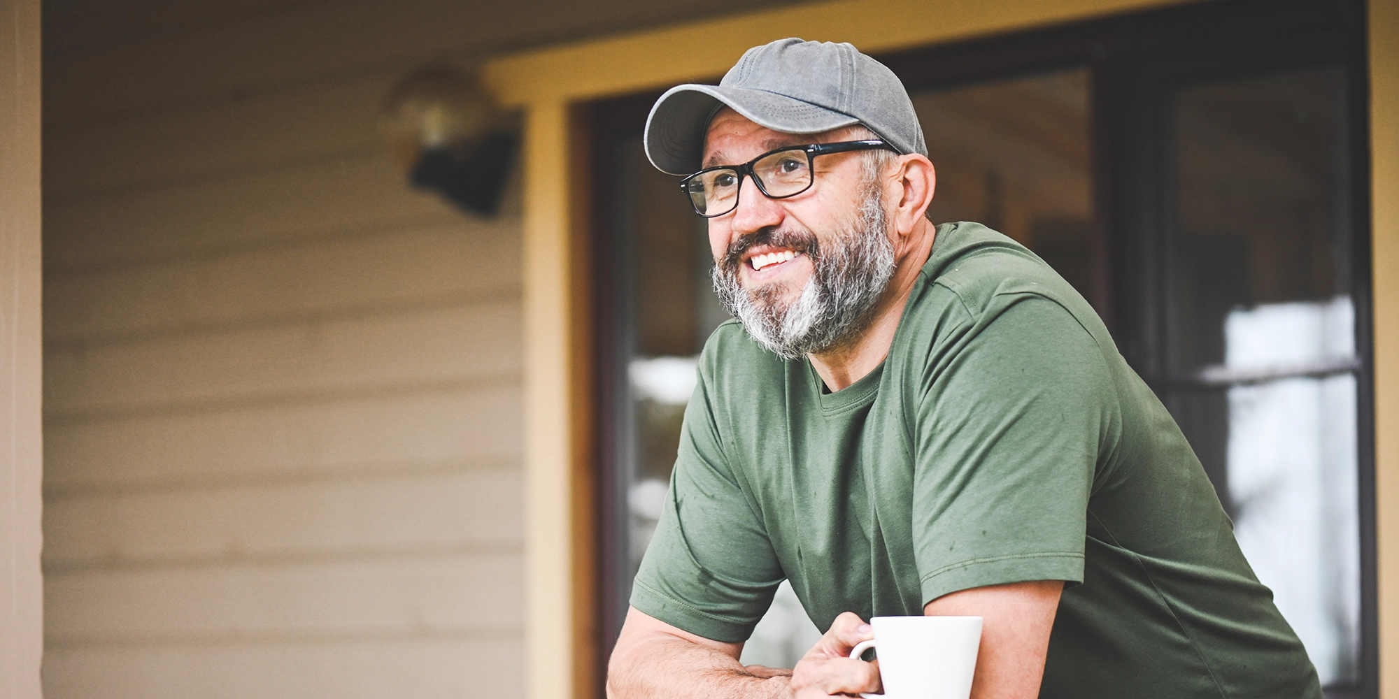 A man leans over a porch railing holding a coffee cup. He happily gazes out into the distance.