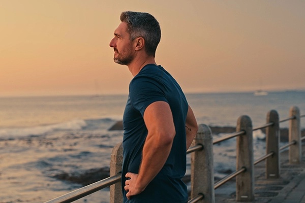 A fit middle-aged man stands on a pier with his hands on his hips. He's peering out thoughtfully over the lake.