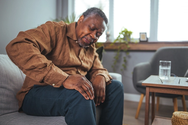 An aging man sitting on a couch winces in pain as he clutches his knee.
