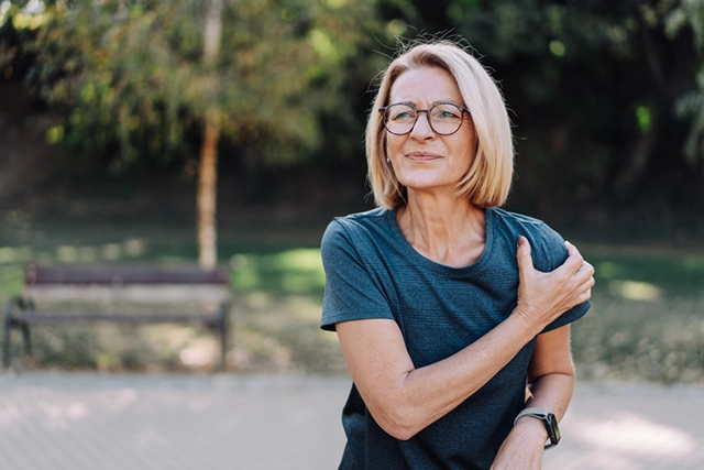 A middle-aged woman walking in a park grabs her shoulder in pain.